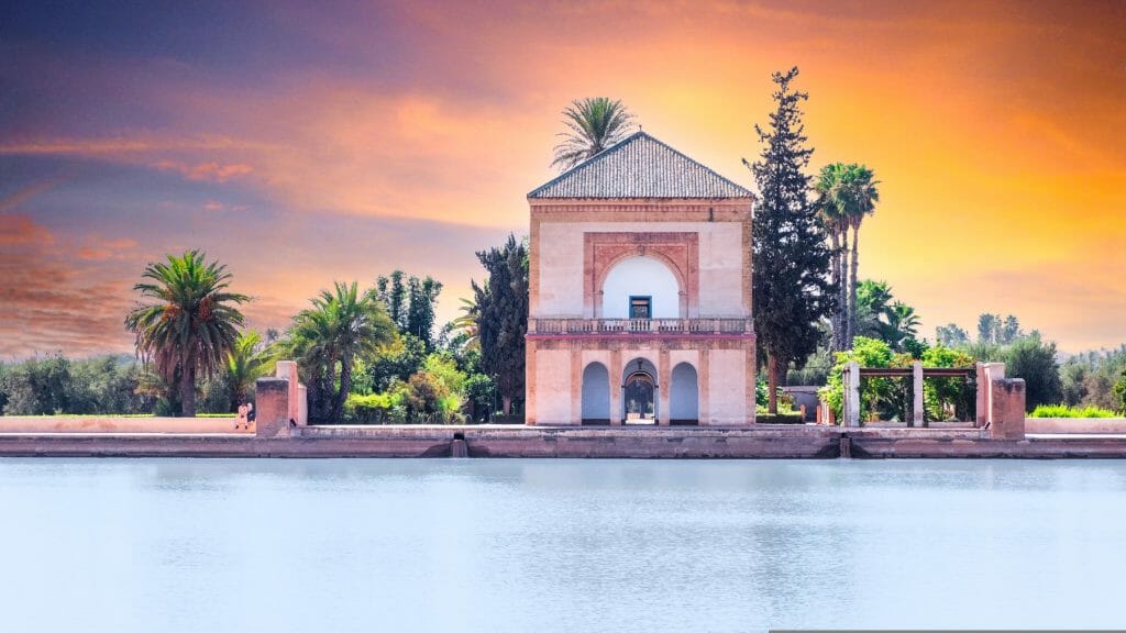 A rectangular building with an arched entrance stands by a calm water basin, surrounded by palm trees, under a vibrant orange and purple sky.